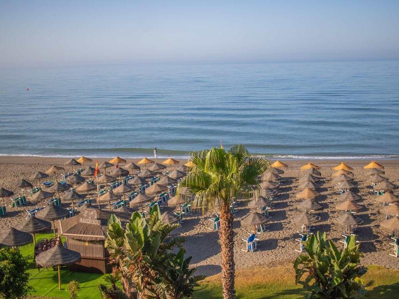 Strand mit Reihen von Sonnenschirmen und Liegen, dahinter ruhiges blaues Meer und klarer Himmel.