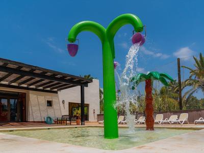 Grüner Wasserspielbereich mit blütenförmigen Duschen neben einem Pool und Sonnenliegen.