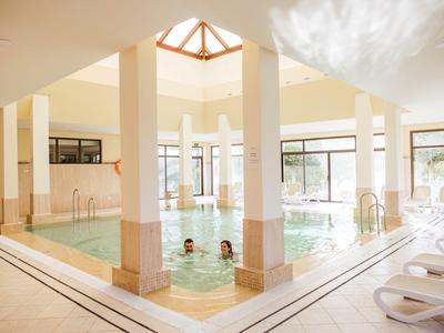 Helles Hallenbad mit Säulen und Liegestühlen, Menschen schwimmen im Wasser.