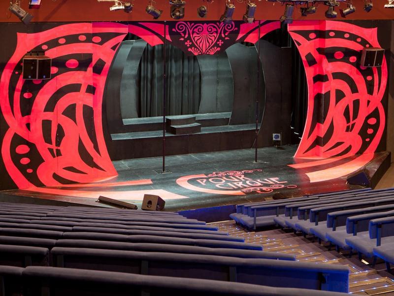 Theaterbühne mit roter abstrakter Wanddekoration und Reihen blauer Sitze im Zuschauerraum.