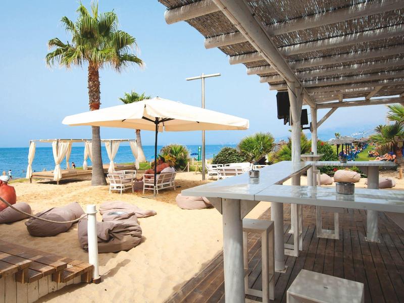 Überdachte Terrasse mit weißen Tischdecken und Korbstühlen in einem Hotelgarten.