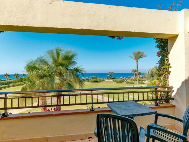Balkon mit Tisch und Stühlen mit Blick auf tropische Pflanzen und das Meer bei klarem Himmel.