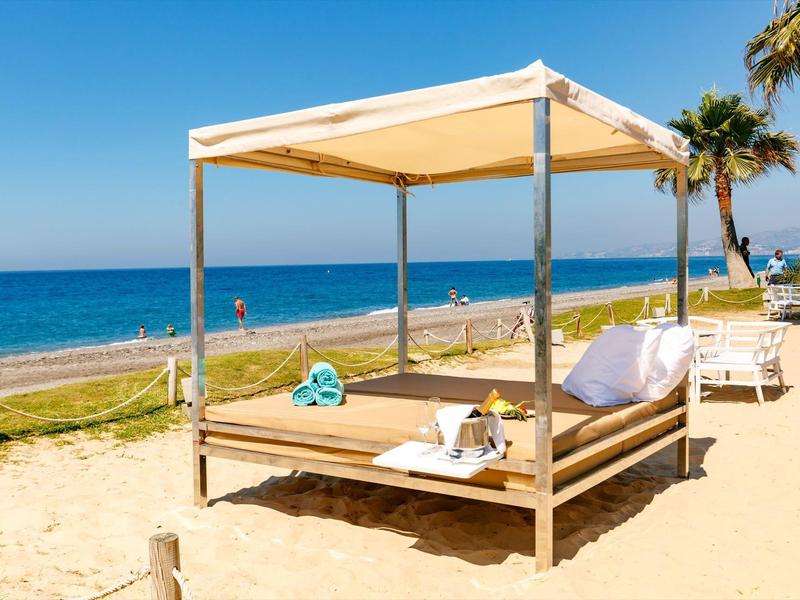Strandliege mit Kissen und Handtüchern unter einem Sonnendach am Sandstrand mit Palmen.
