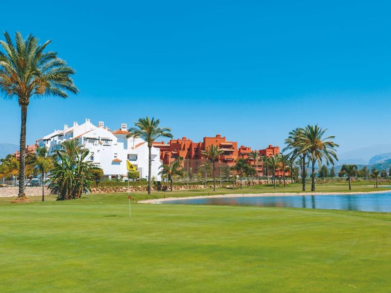 Grüner Golfplatz mit Palmen und einem Hotelgebäude im Hintergrund bei klarem Himmel.