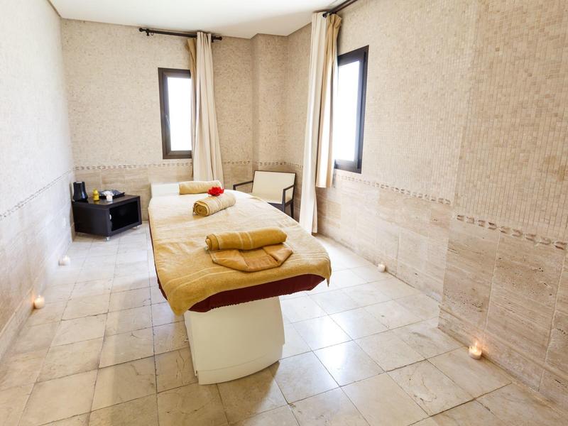 Hell beleuchteter Wellnessraum mit Massageliege, Handtüchern und Kerzen auf Fliesenboden.