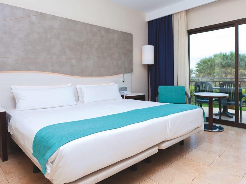 Helles Hotelzimmer mit großem Bett, türkisfarbenem Läufer und Balkon mit Blick ins Grüne.