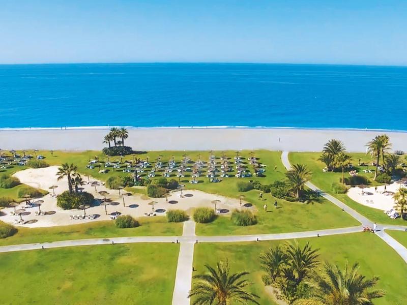 Weitläufige Strandanlage mit Liegestühlen, Sonnenschirmen und einem großen Pool am Meer.