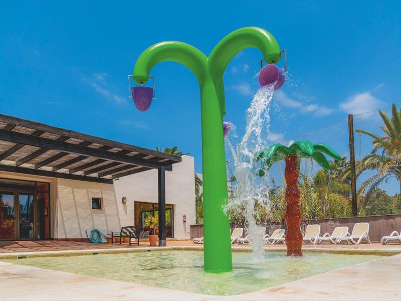 Bunter Wasserspielplatz mit grünen und braunen Pflanzenformen neben einem Pool unter blauem Himmel.