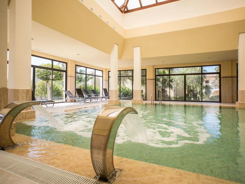 Helles Hallenbad mit Wasserfällen, großen Fenstern und bequemen Liegestühlen.