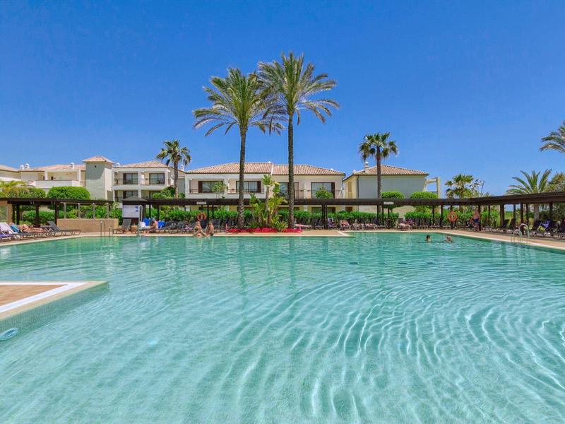 Großer Hotelpool mit Palmen und Einfamilienhäusern im Hintergrund an einem sonnigen Tag.