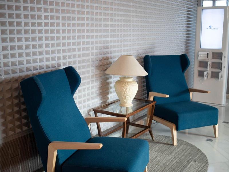 Zwei blaue Sessel mit Holzrahmen neben einem kleinen Tisch mit einer Lampe vor einer weißen Wand.