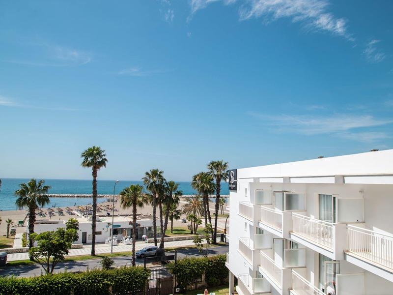 Hotelzimmer mit Balkon und Meerblick am Strand mit Palmen unter blauem Himmel.