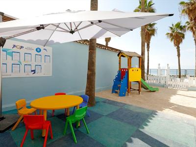 Kinderspielplatz mit bunten Stühlen und Sonnenschirm neben Klettergerüst und Rutsche.