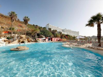 Großer Pool mit klarem Wasser, umgeben von Liegestühlen und Palmen vor einem Hotel.