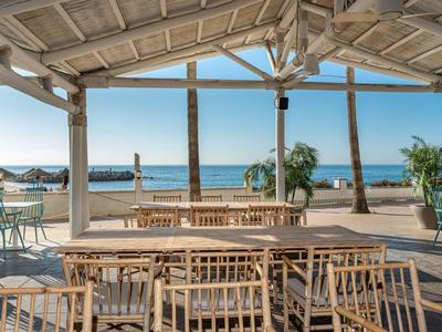 Offener Pavillon mit Holzmöbeln am Strand mit Palmen und Meer im Hintergrund.
