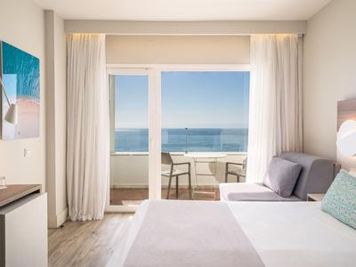 Helles Hotelzimmer mit Doppelbett und Balkon mit Meerblick bei klarem Himmel.