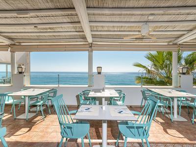 Terrasse mit türkisfarbenen Stühlen und Tischen mit Blick auf das Meer bei klarem Himmel.