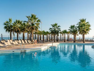 Ein ruhiger Pool mit Liegestühlen und Palmen am Meer an einem sonnigen Tag.