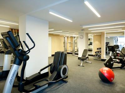 Moderner Fitnessraum mit Trainingsgeräten, hellem Boden und neutraler Wandfarbe.