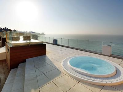 Moderne Dachterrasse mit rundem Whirlpool, Glasgeländer und Blick auf ruhiges Meer bei Sonnenaufgang.