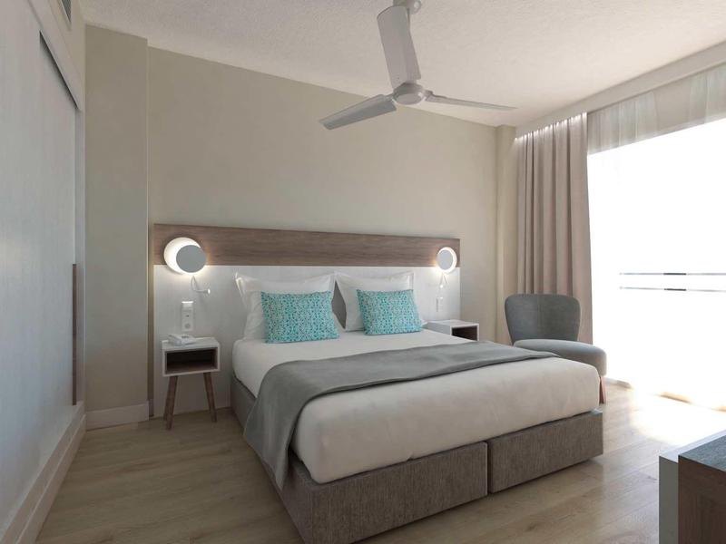 Hell eingerichtetes Schlafzimmer mit Doppelbett, Deckenventilator, Sessel und großem Fenster mit Vorhängen.