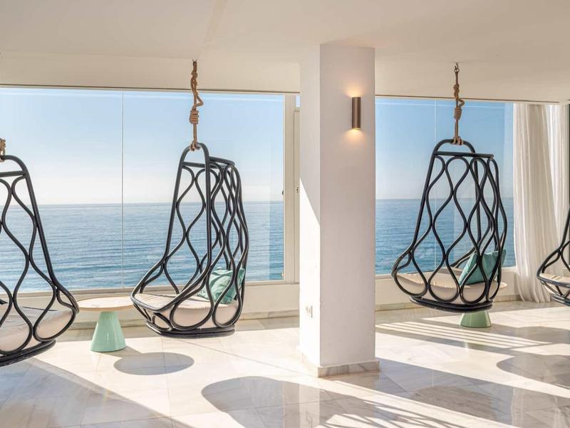 Vier schwarze Hängesessel aus Metall hängen vor großen Fenstern mit Meerblick in einem hellen Raum.