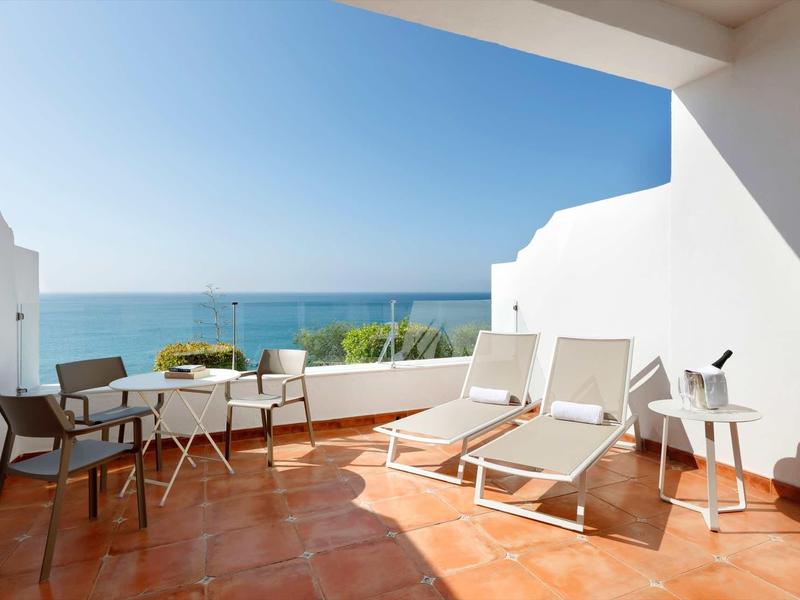 Terrasse mit zwei Liegen, vier Stühlen, Tischen und Blick auf das blaue Meer unter klarem Himmel.