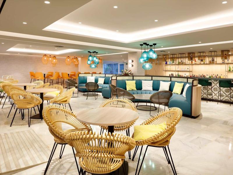 Moderner Gastraum mit hellen Wänden, Rattanstühlen und blauen Polstern, warm beleuchtet.