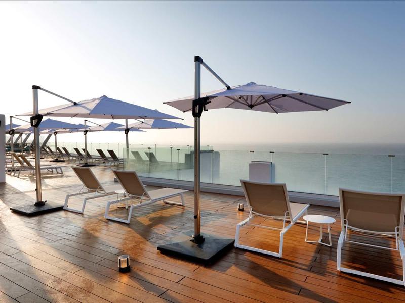 Holzterrasse mit Liegestühlen und großen Sonnenschirmen am ruhigen Meer bei klarem Himmel.