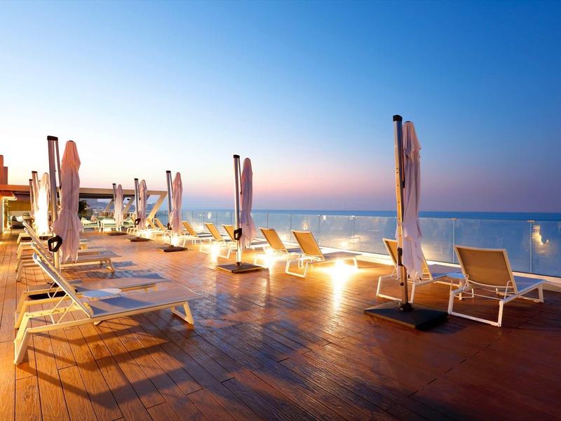 Poolterrasse mit Liegestühlen und geschlossenen Sonnenschirmen bei Sonnenuntergang am Meer.