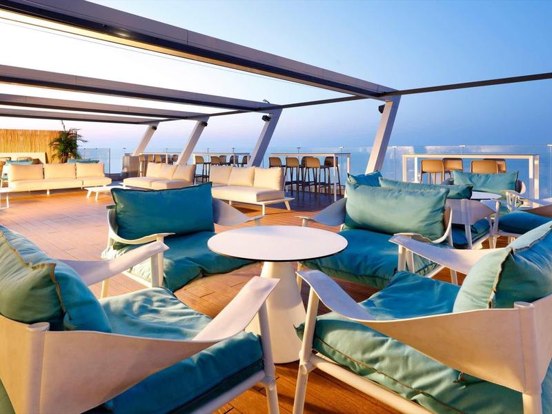 Offene Terrasse mit blauen Sitzpolstern, weißen Tischen und Blick auf Meer und Himmel bei Sonnenuntergang.