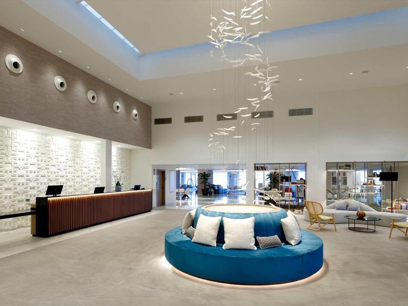 Moderne Hotellobby mit rundem, blauem Sofa, Rezeption links und großer Glaswand im Hintergrund.