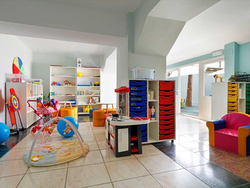 Bunter, heller Kindergartenraum mit Spielzeug, Sitzgelegenheiten und regalen auf Kachelboden.