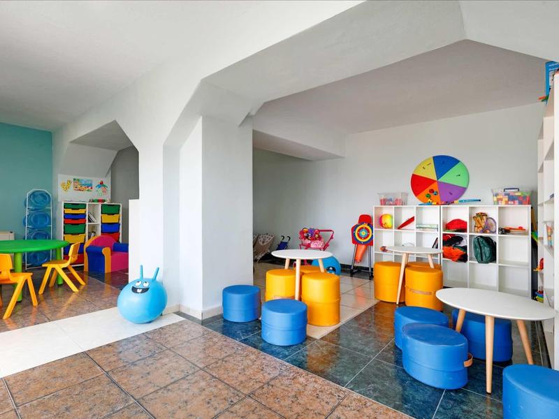 Buntes Kinderspielzimmer mit Tischen, Stühlen, Sitzhockern und Spielzeug in zwei Bereichen.