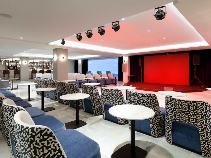 Moderner Veranstaltungsraum mit blauen Sitzen, weißen Tischen und roter Bühne im Hintergrund.