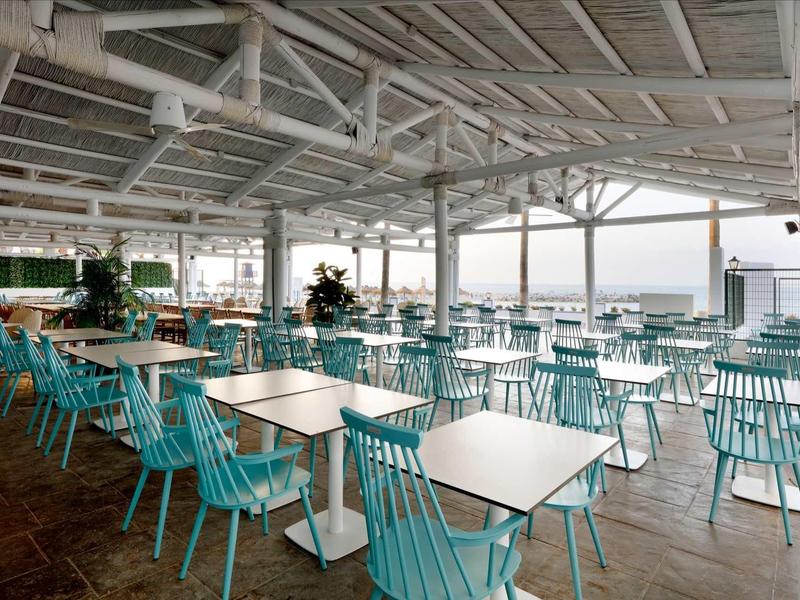 Große, offene Terrasse mit türkisfarbenen Stühlen und weißen Tischen unter einem weißen Dach.