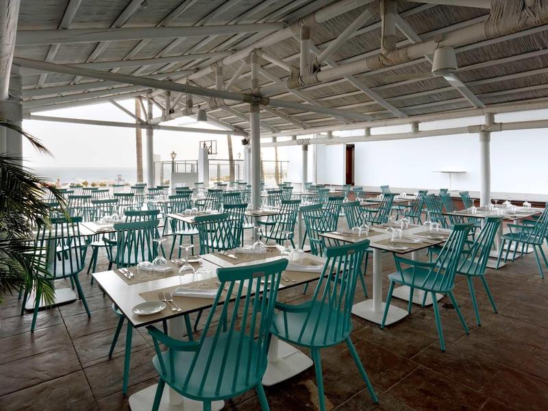 Großer, heller Gastraum mit vielen türkisfarbenen Stühlen und gedeckten Tischen, offener Meerblick.