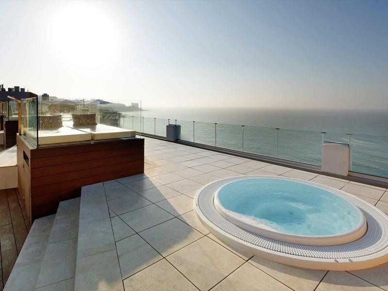 Moderne Dachterrasse mit rundem Whirlpool, Glasgeländer und Blick auf ruhiges Meer bei Sonnenaufgang.