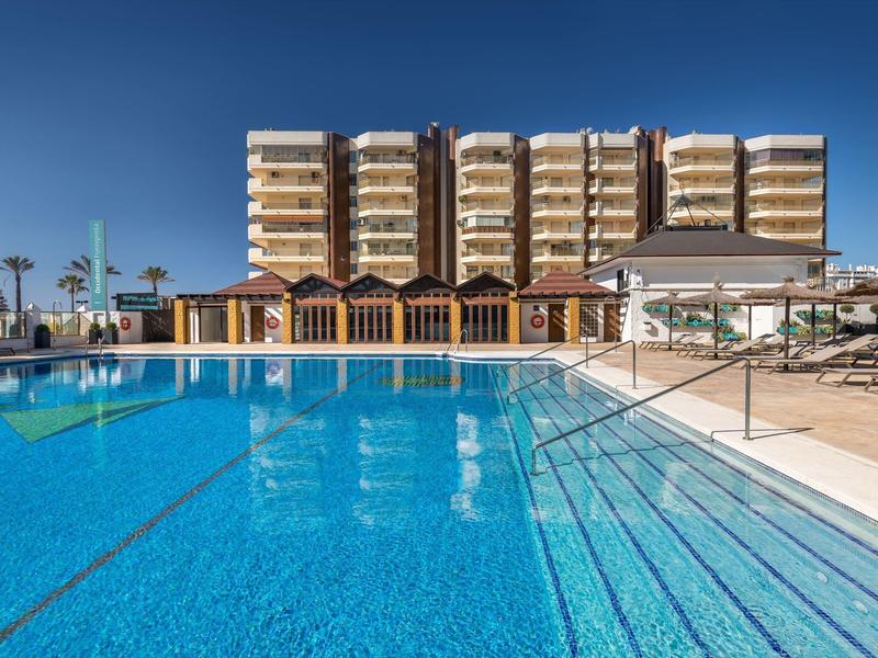 Großer blauer Pool mit Treppen, Liegestühlen und mehrstöckigem Gebäude im Hintergrund.