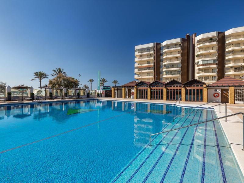 Großer Außenpool mit klarem Wasser vor mehrstöckigem Hotel und Palmen unter blauem Himmel.