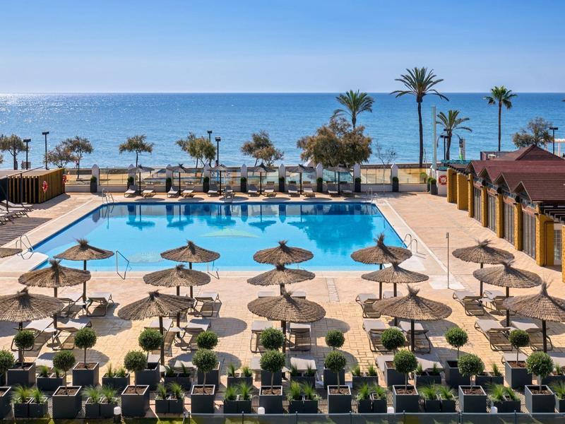 Großer Swimmingpool mit Sonnenschirmen, Palmen und Blick auf das blaue Meer bei klarem Himmel.