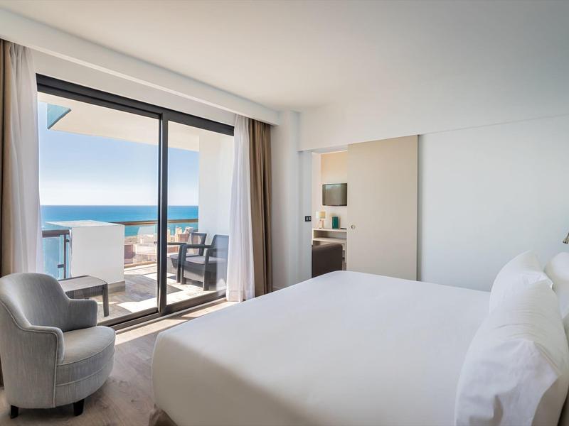 Helles Schlafzimmer mit großem Bett, Sessel und Meerblick durch bodentiefe Fenster und Terrasse.