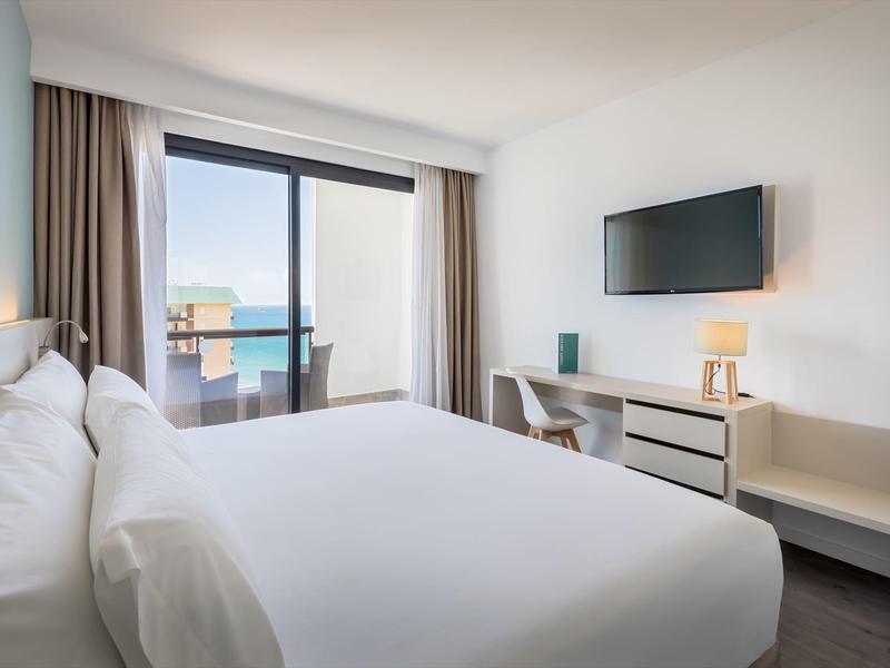 Helles Schlafzimmer mit großem Bett, Fernsehbildschirm, Schreibtisch und Meeresblick durch große Fenster.