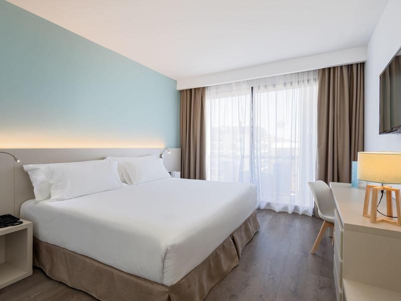 Modernes Hotelzimmer mit großem Doppelbett, blauer Akzentwand, Schreibtisch und großem Fenster mit Vorhängen.