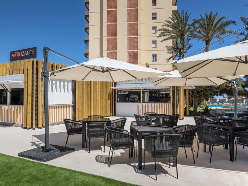 Moderner Außenbereich mit schwarzen Stühlen, Tischen, weißen Sonnenschirmen und Palmen im Hintergrund.