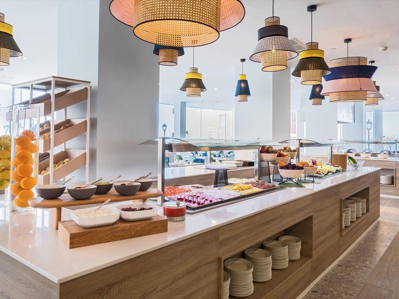 Buffet mit vielfältigen Speisen auf hellem Holz, große Fenster und bunte Hängelampen im Raum.