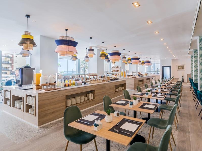 Modernes Restaurant mit Holztischen, grünen Stühlen und bunten Designerlampen an der Decke.