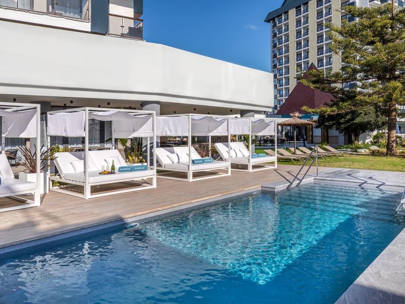 Moderner Außenpool mit weißen Liegen und blauen Kissen neben Holzterrasse und Hochhaus im Hintergrund.