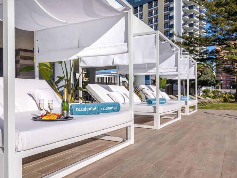 Weiße Lounge-Betten mit farbigen Kissen stehen auf Terrasse neben Hochhaus und Bäumen bei Sonnenschein.