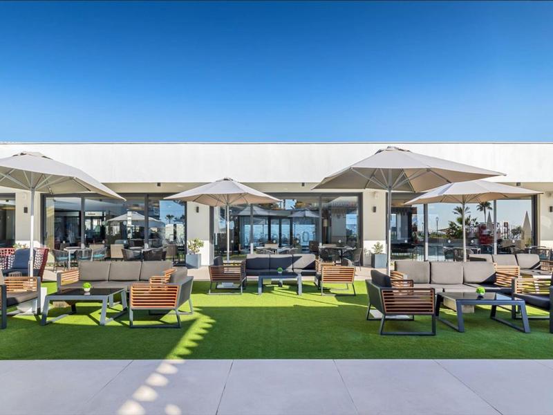 Moderner Außenbereich mit Sitzgruppen, Sonnenschirmen und großem Glasgebäude bei blauem Himmel.